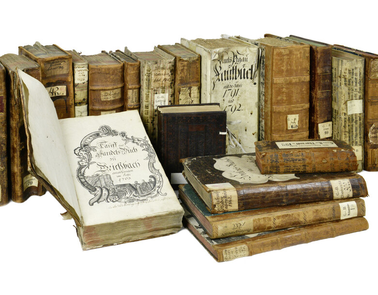 Das Foto zeigt mehrere alte in Leder eingebundene Bücher, wovon einige in Reihe stehen, andere gestapelt liegen und eines aufgeschlagen ist.