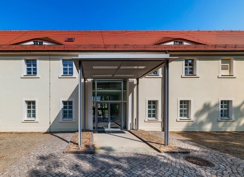 Außenansicht des Schlossgebäudes mit überdachtem Eingang vor, helle mit Fenster untergliederte Fassade mit roten Dachziegeln und Gauben vor blauen Himmel, im Vordergrund gepflasteter Weg